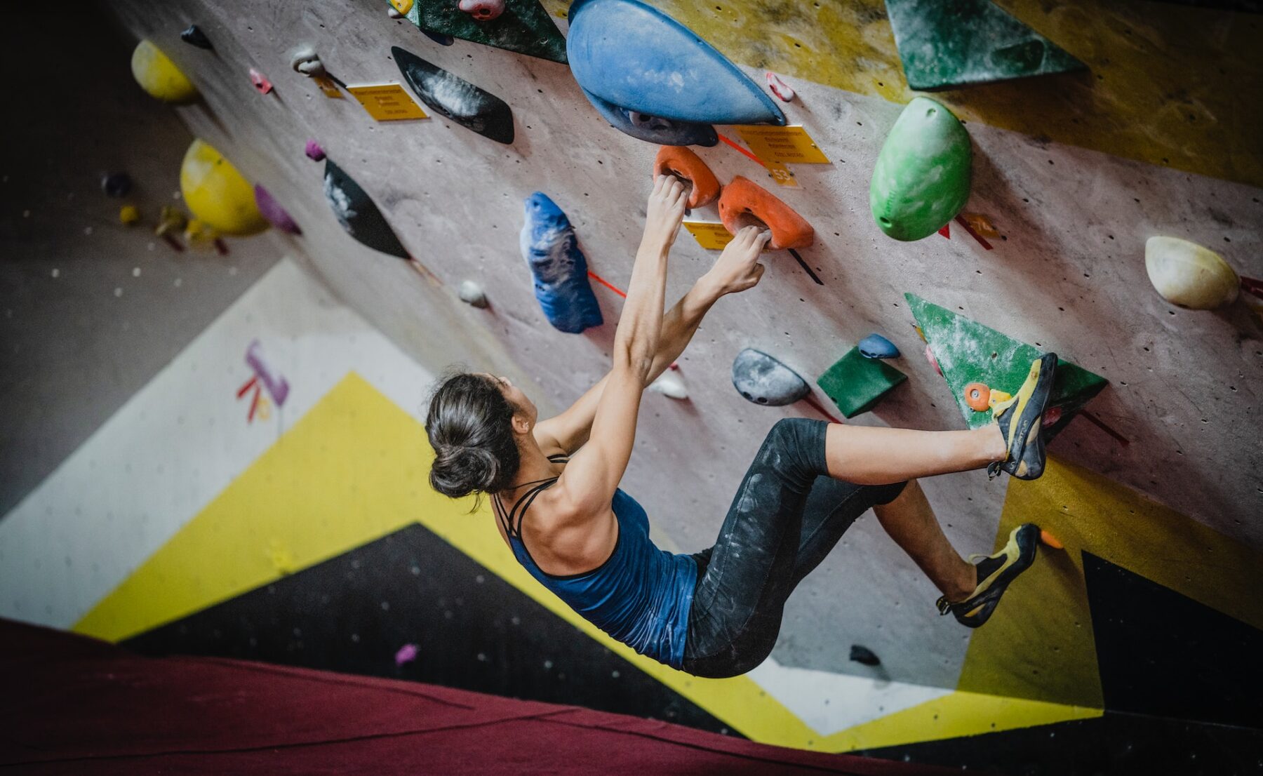A person in a blue tank top and black pants is climbing an indoor bouldering wall with various colorful holds. The ceiling and walls are marked by angled sections, creating a challenging route. The red floor below provides a safety mat.
