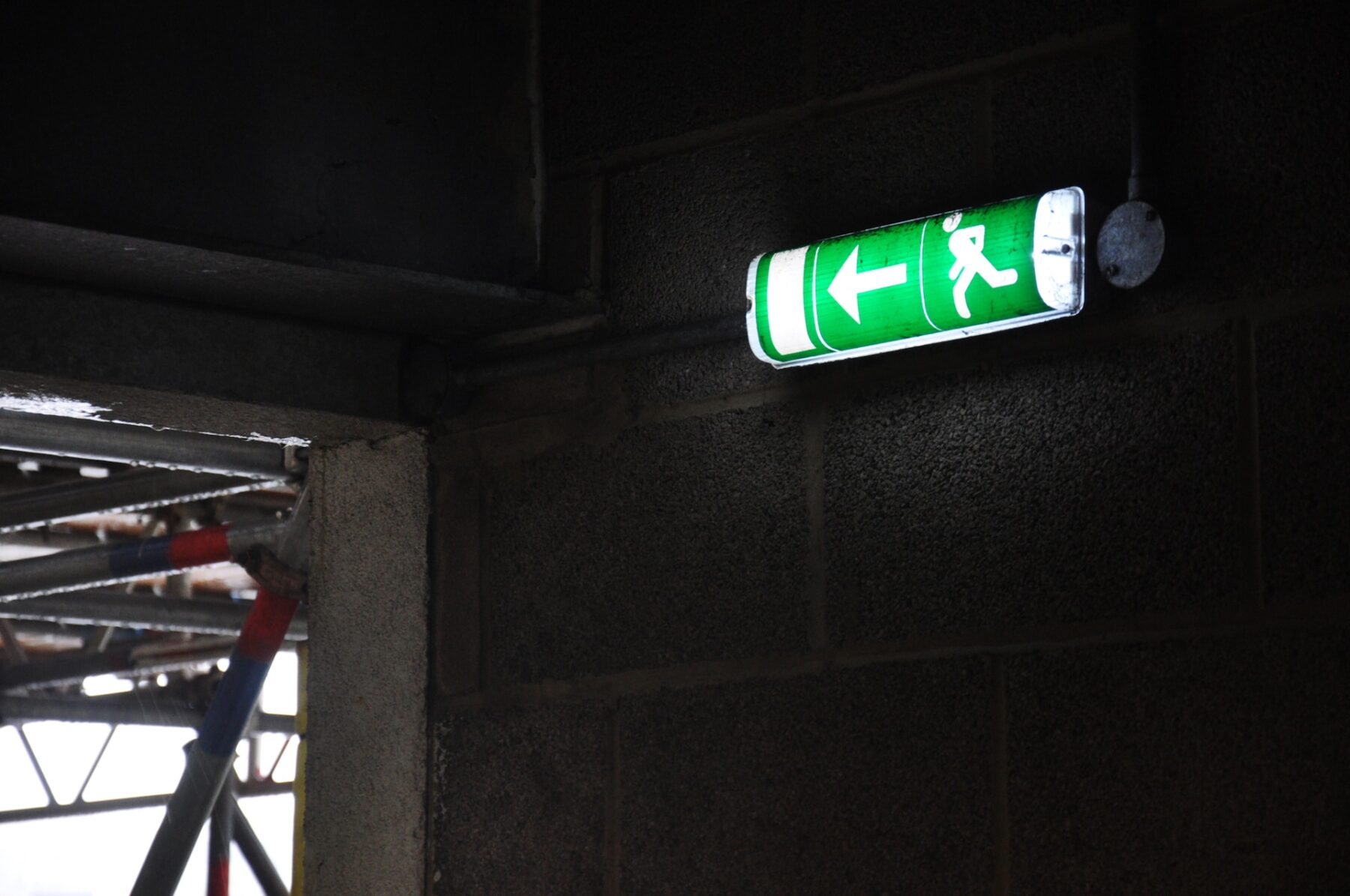 A green exit sign mounted on the side of a building, featuring bold white lettering that is partially obscured by shadows.